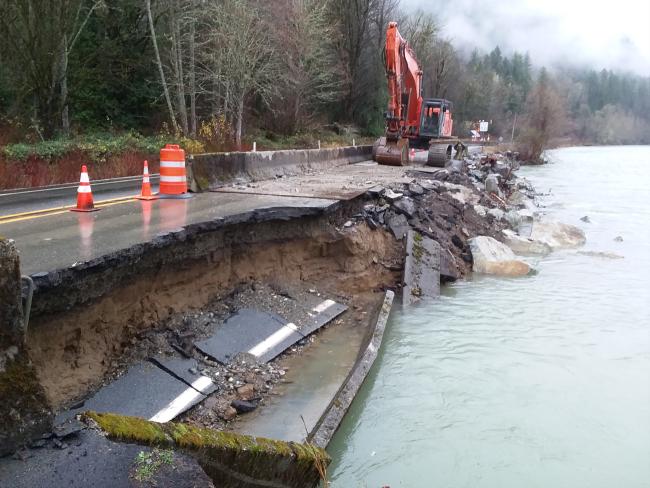 An excavator reconstructs the SR 20 highway embankment along the Skagit River following a November 2017 flood. The eastbound lane is closed and a section of the road is crumbling into the river channel.