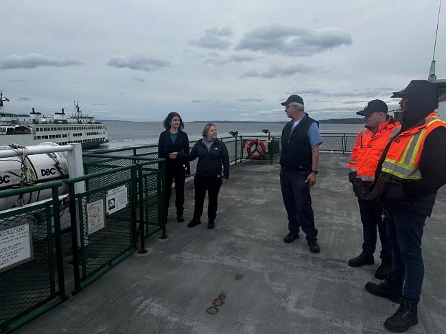 Five people talking on the outdoor deck of a ferry with another ferry in the water in the background